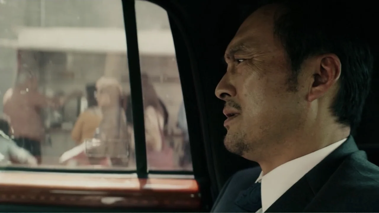 Bel Canto (2018) movie still: medium — A man in a suit sits inside a car looking out a window. Blurred figures of pedestrians are visible on the…