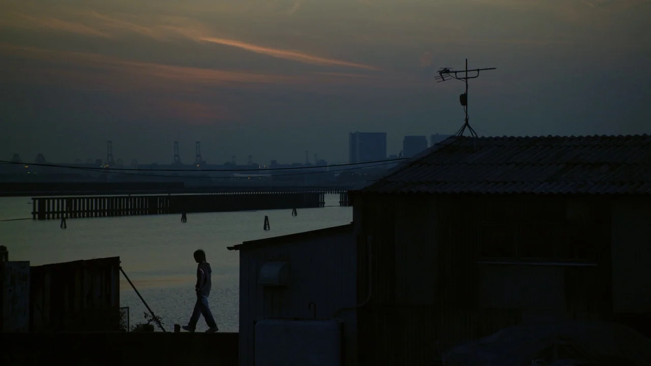 Confessions (2010) movie still: wide — An industrial waterfront area with metal structures and a distant city skyline at dusk. A lone person walks…
