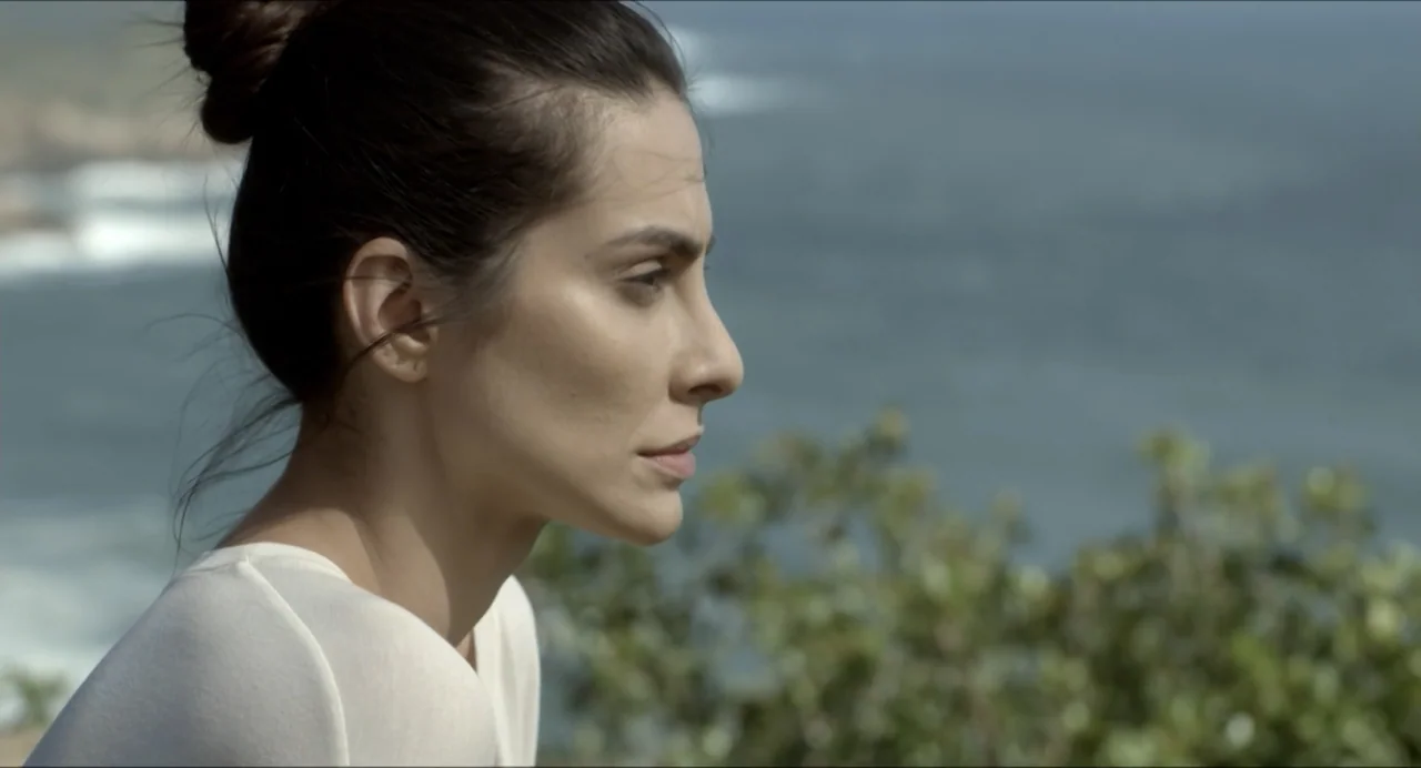 Suspension Points (2022) movie still: closeup — A woman stands against a backdrop of a blurred ocean and coastal foliage. She looks toward the…