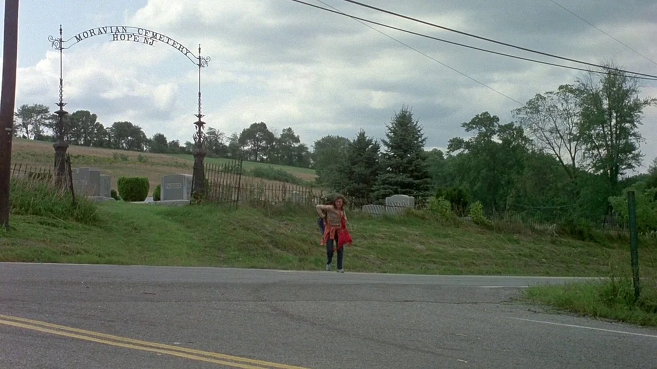 Friday the 13th (1980) movie still: wide — A rural road passes by an iron cemetery gate under an overcast sky. A young person with a backpack…