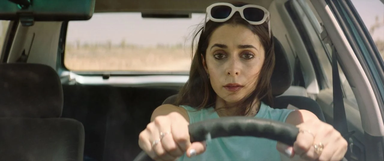 Palm Springs (2020) movie still: medium — A woman sits in the driver seat of a car during the day. She grips the steering wheel with both hands…