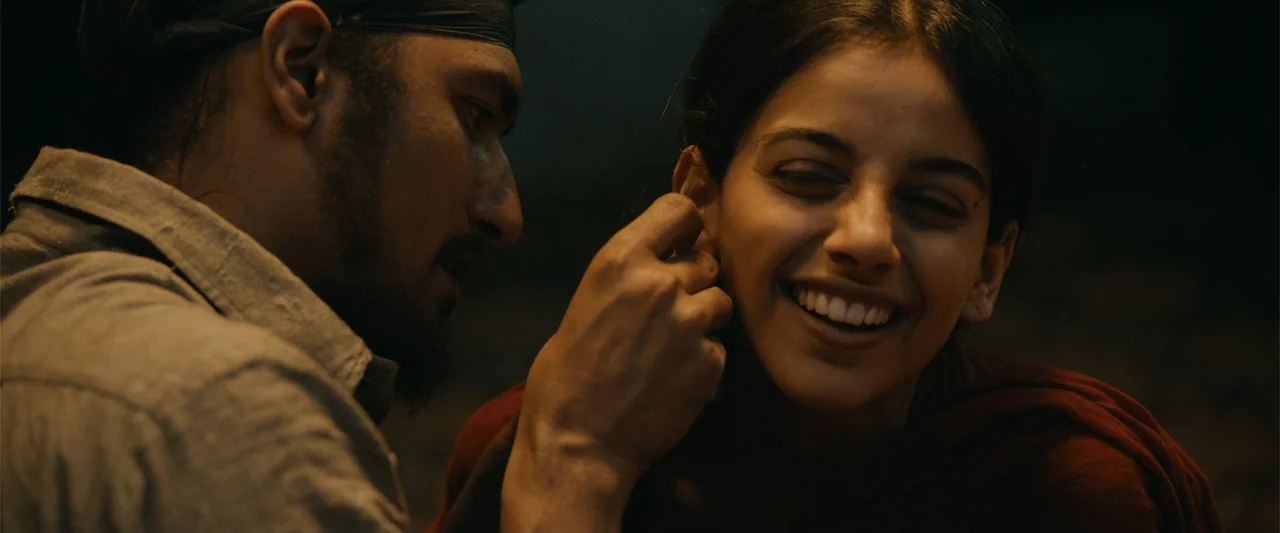 Sardar Udham (2021) movie still: medium — A dimly lit interior space with dark walls and minimal background detail. A man stands close to a woman…