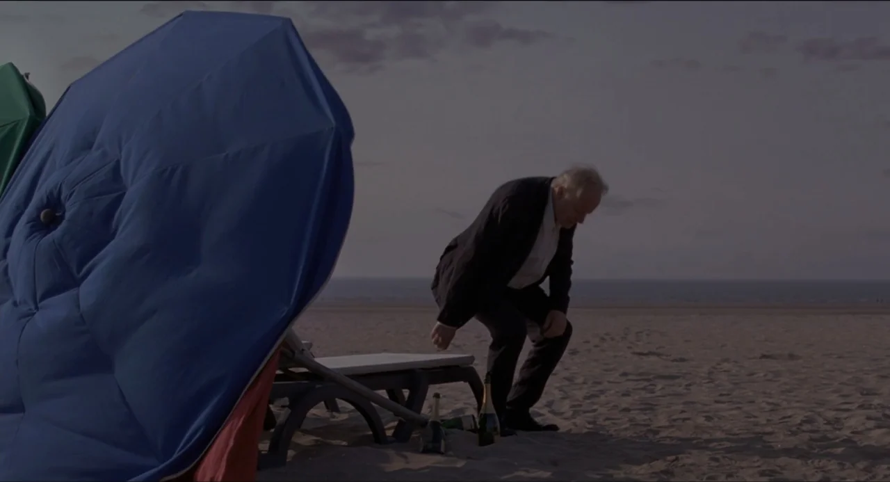 Sentimental Value (2025) movie still: medium — An empty beach at dusk features a single large blue umbrella near a lounger. A mature man in formal…