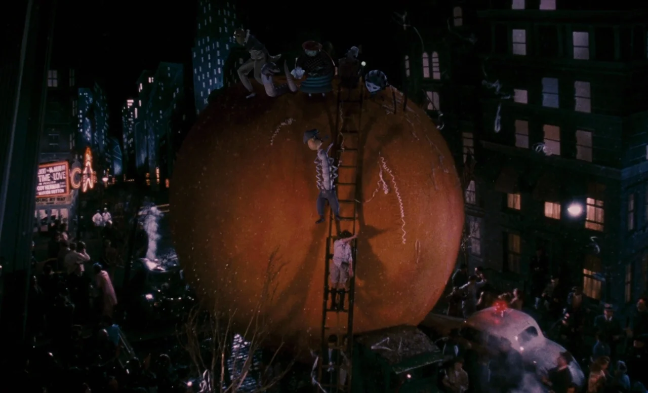 James and the Giant Peach (1996) movie still: wide — A massive orange sits in the middle of a nighttime city street filled with onlookers. Several…