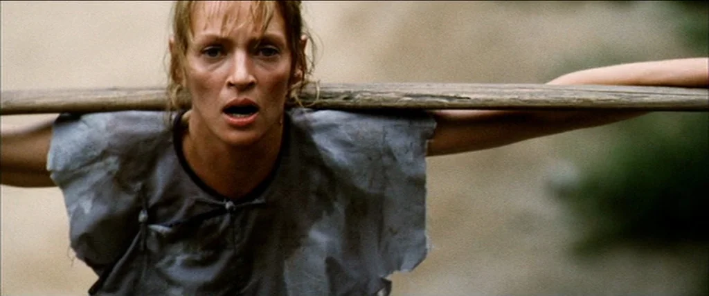 Kill Bill Vol. 2 (2004) movie still: medium — A woman stands in a forest area with a large piece of wood. She has a wide-eyed expression while…