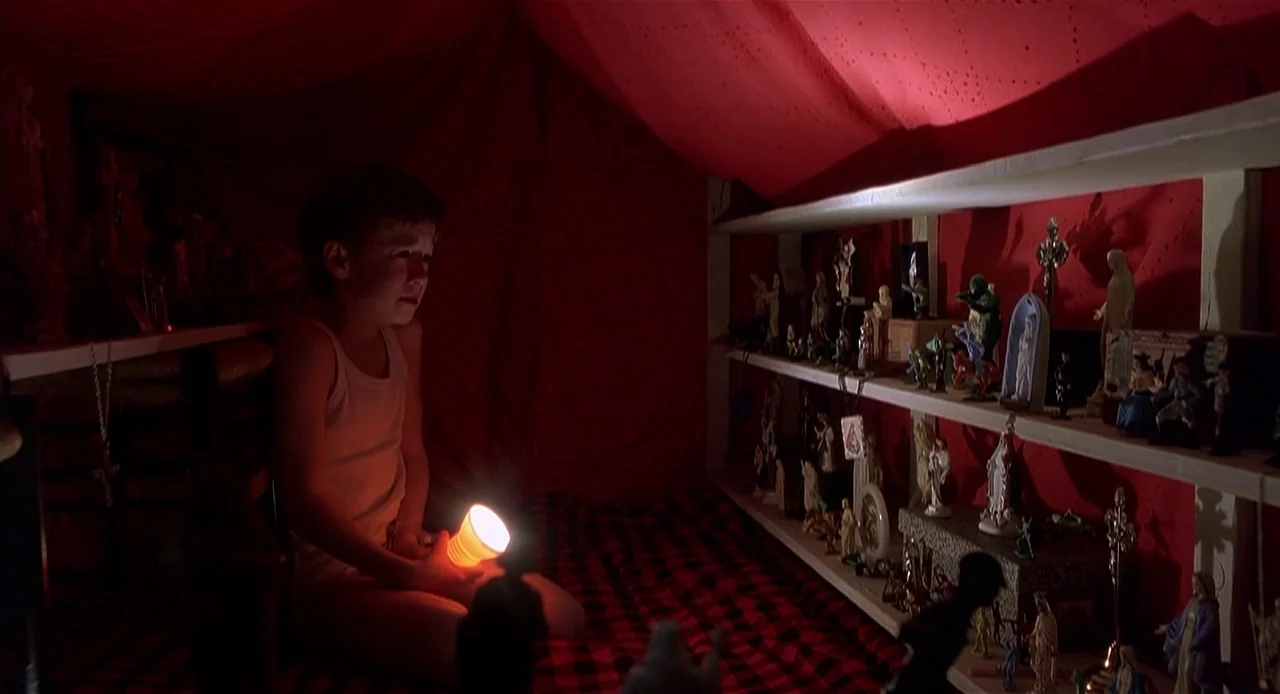 The Sixth Sense (1999) movie still: medium — A dark tent interior is lined with red fabric and shelves filled with figurines. A young boy sits on a…