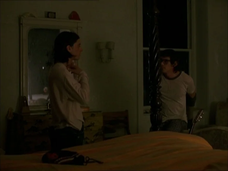 Last Days (2005) movie still: medium — A dimly lit room with dark walls and a four-poster bed frame. One man adjusts his shirt near a dresser while…