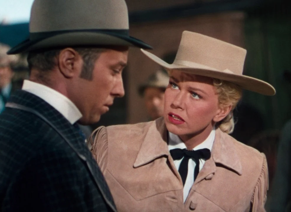 Calamity Jane (1953) movie still: medium — An outdoor western street setting with blurred wooden structures in the background. A woman in a tan…