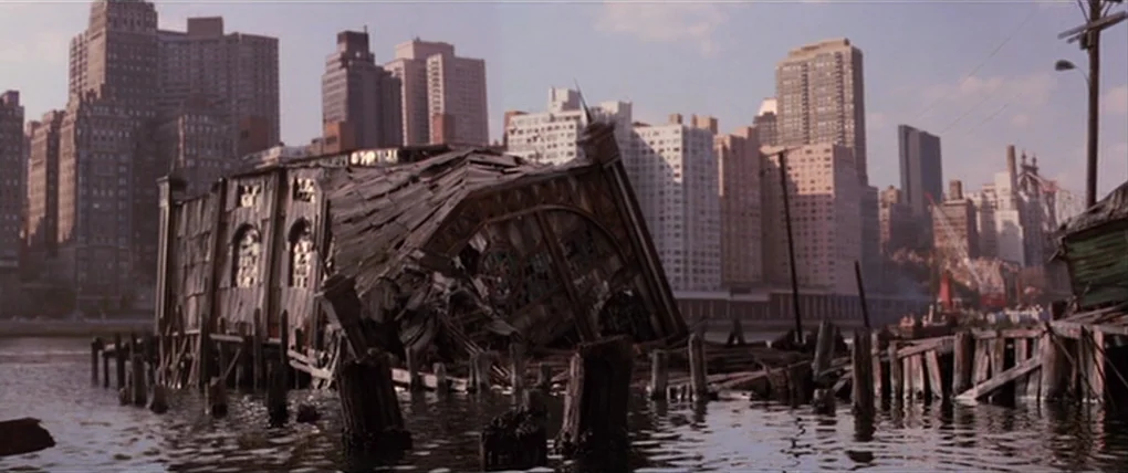 Spider-Man 2 (2004) movie still: establishing — A dilapidated wooden structure sags into the water along a developed city shoreline. Tall…