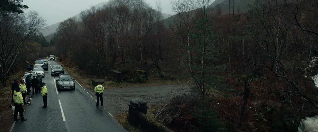 My Son (2021) movie still: wide — A rural asphalt road cuts through a hilly area with sparse trees and brush. Several police vehicles are parked on…