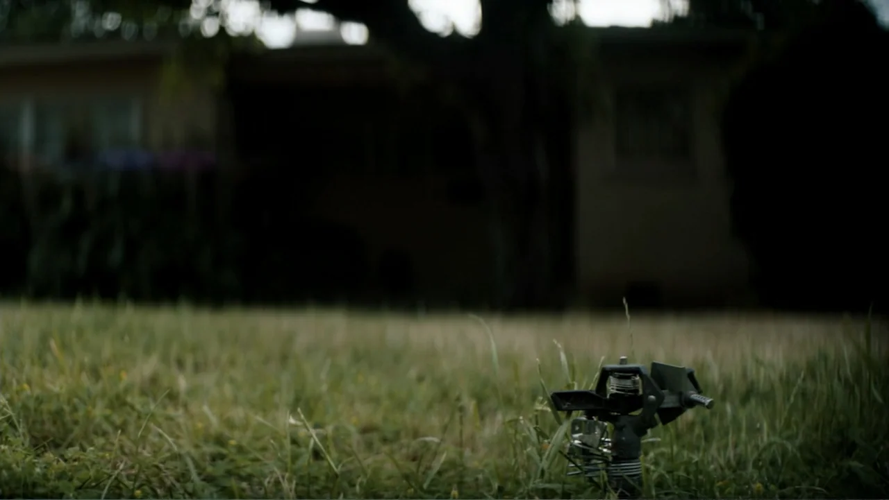 Nightingale (2014) movie still: medium — An outdoor suburban lawn is covered with grass and a metal sprinkler. The sprinkler stands in the…