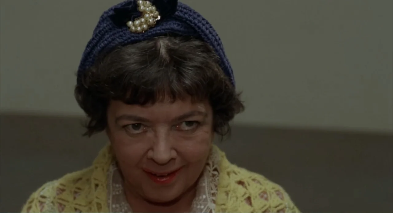 Cottonpickin' Chickenpickers (1967) movie still: closeup — A plain interior room with a neutral grey wall as a backdrop. An older woman with dark…