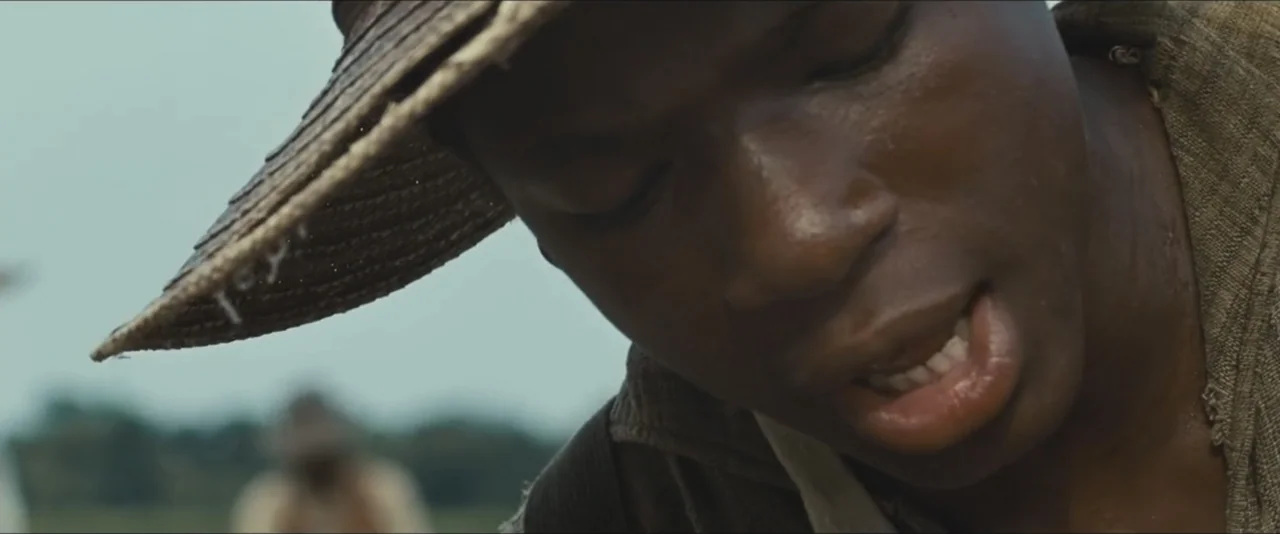 12 Years a Slave (2013) movie still: closeup — An open rural field under a bright daylight sky. A young man wearing a straw hat looks downward while…