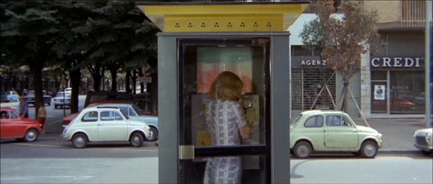 Four Flies On Grey Velvet (1971) movie still: medium — A street corner features a yellow-topped metal telephone booth centered in the frame. A woman…