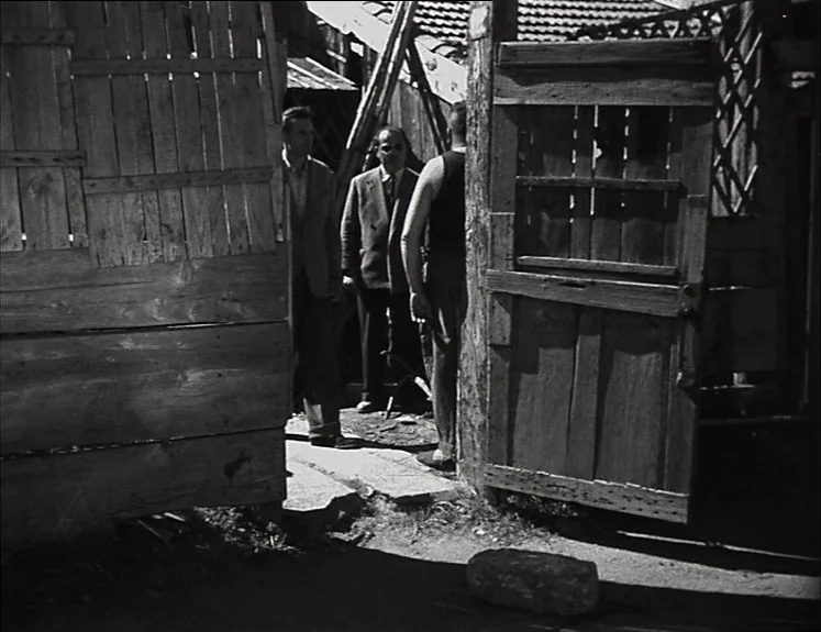 La Pointe Courte (1955) movie still: medium — A rustic enclosure features weathered wooden walls and a worn gate with lattice detail. Three men in…