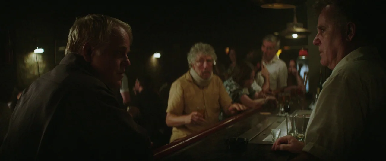 God’s Pocket (2014) movie still: medium — A dimly lit bar contains wooden furniture and patrons sitting in the shadows. A man at the counter listens…
