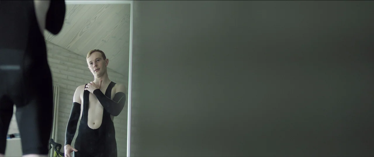 The Feast (2021) movie still: medium — A man stands before a large wall mirror inside a room with light-colored brick walls. The reflection reveals…