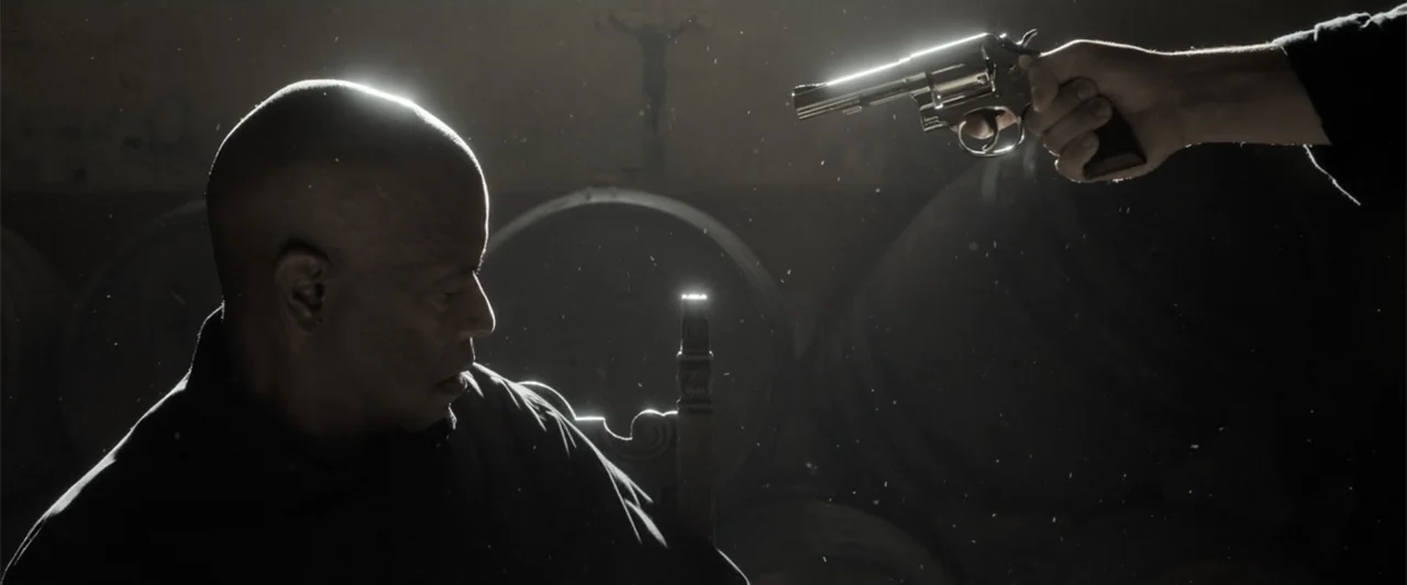 The Equalizer 3 (2023) movie still: medium — A dark room contains stacked wooden barrels and a faint crucifix on the back wall. An older man sits in…