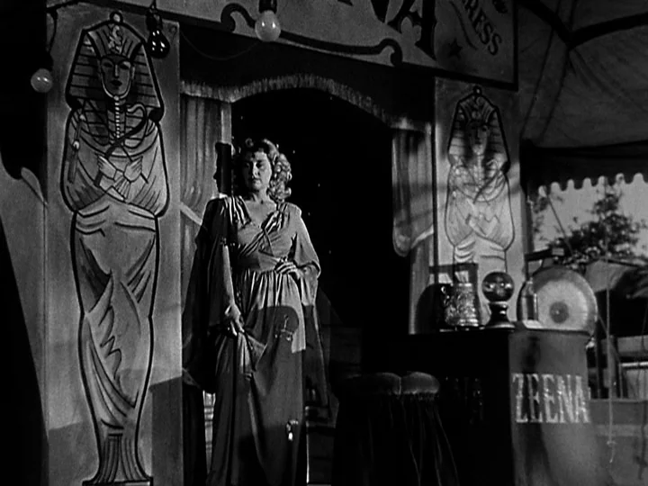 Nightmare Alley (1947) movie still: fullbody — A carnival fortune teller booth features painted Egyptian-style figures on flanking boards. A woman…