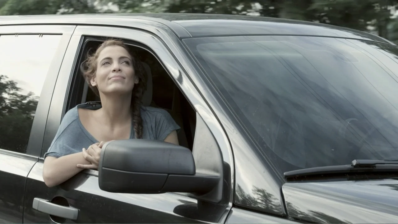 Locas y atrapadas (2014) movie still: medium — A dark SUV travels along a road with a woman hanging out the window. The woman rests her folded arms…