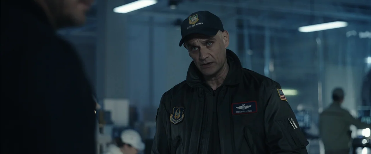 Moonfall (2022) movie still: medium — A spacious industrial hangar features rows of blurred equipment and overhead lighting fixtures. A middle-aged…