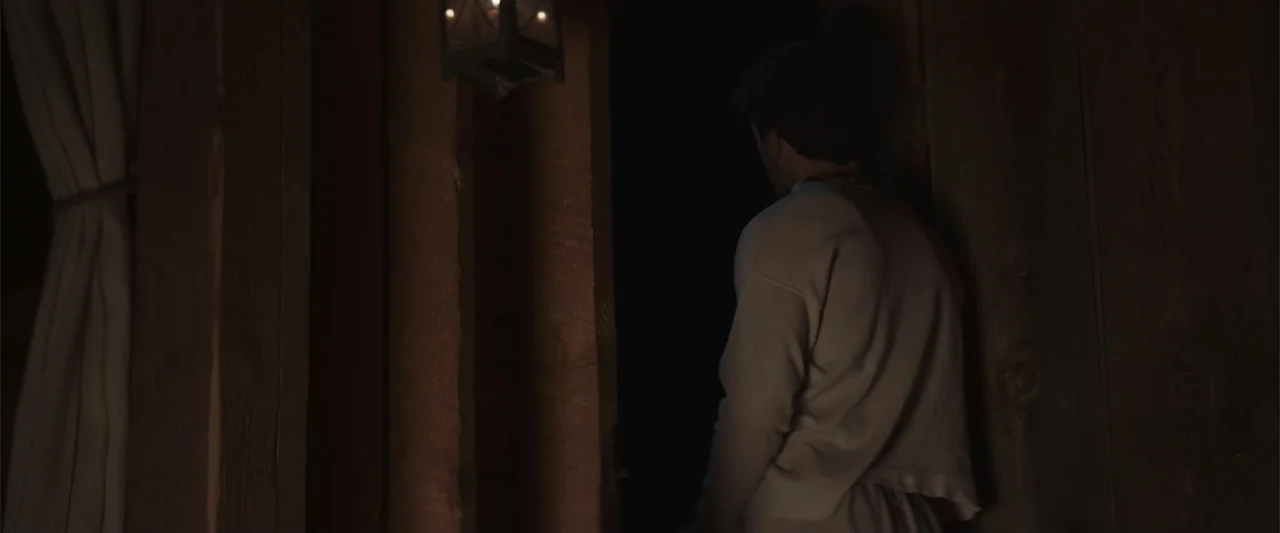 The Wind (2005) movie still: medium — An interior space features rough wooden walls and a dark open doorway. A man stands at the threshold looking…