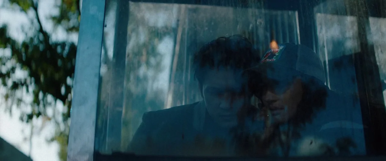 Broadcast Signal Intrusion (2021) movie still: medium — Two people are visible through a reflective glass partition in an outdoor setting. The…