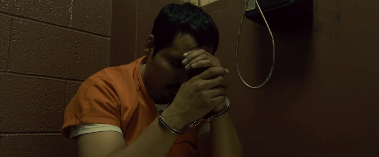 Frontera (2014) movie still: medium — A small room features bare painted block walls and a wall-mounted telephone fixture. A man in an orange…