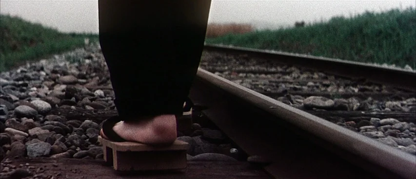 Jigoku (1960) movie still: closeup — A pair of wooden geta sandals stand on gravel beside rusty train tracks. The person wearing the sandals stands…
