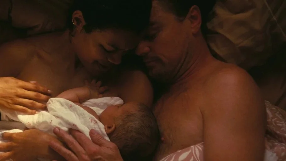 One Battle After Another (2025) movie still: closeup — A close-up of a man, woman, and their newborn baby lying together in bed. The parents look…