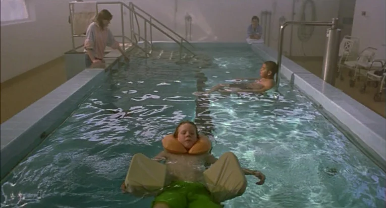 A Room For Romeo Brass (1999) movie still: wide — A clinical tiled pool with metal railings and medical equipment in the background. A boy floats in…