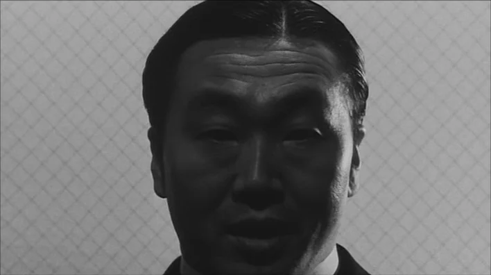 Death By Hanging (1968) movie still: closeup — A man's face is illuminated in high-contrast chiaroscuro lighting against a simple textured…