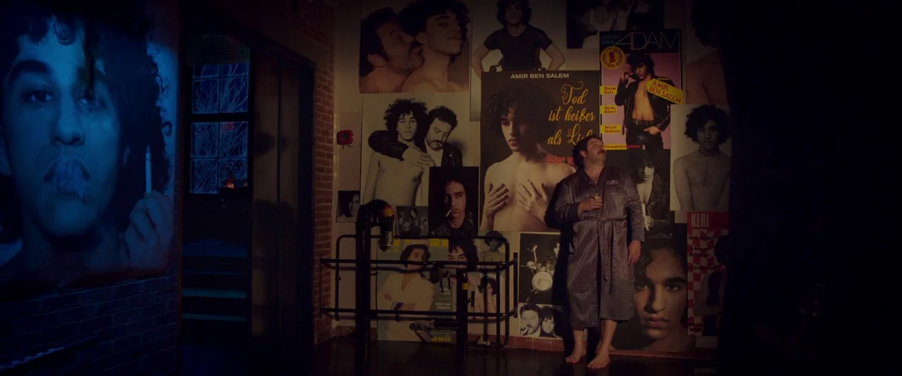 Peter Von Kant (2022) movie still: wide — An interior space features a wall densely covered with various oversized posters of men. A man wearing a…