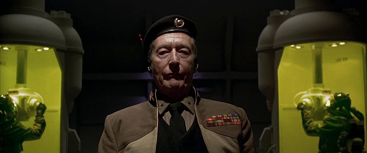 The Fifth Element (1997) movie still: medium — An elderly military leader stands in a dark, tech-focused room flanked by green-lit pods. Two figures…