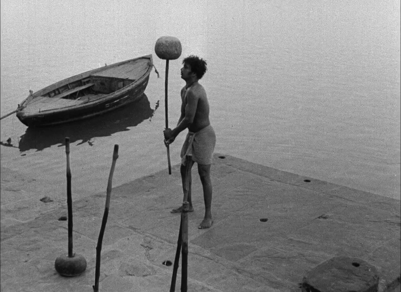 Aparajito (1956) movie still: fullbody — A stone riverside landing has several weighted wooden poles placed on the ground. A man holds one of the…