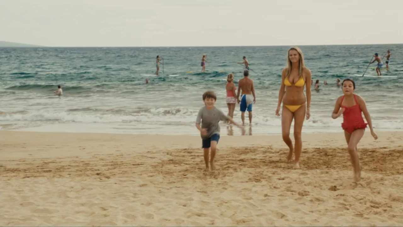 Just Go with It (2011) movie still: fullbody — A sunny sand beach with several people in the background swimming and paddle boarding. A woman and…