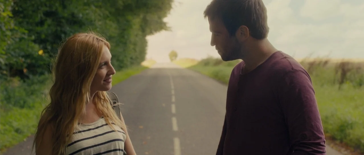 Road Games (2015) movie still: medium — An asphalt road stretches into the distance between grassy verges and dense green foliage. A man and a woman…