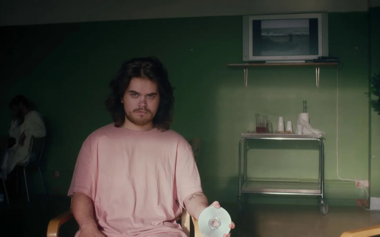 Knocking (2021) movie still: medium — A man with long hair sits in front of a dull green wall in a room. He holds a shiny compact disc in his hand…