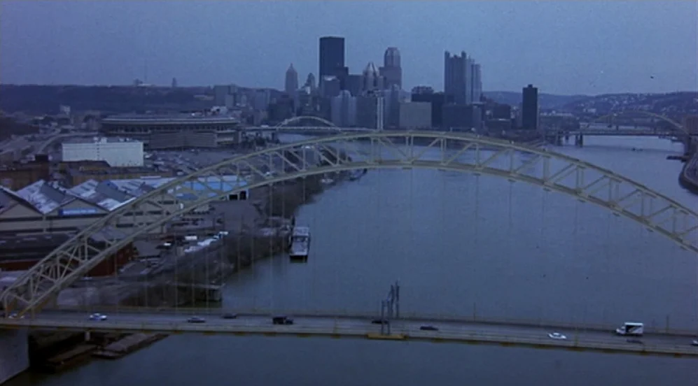 Groundhog Day (1993) movie still: establishing — A wide view shows a large arched bridge crossing a river toward a city skyline. The cityscape…