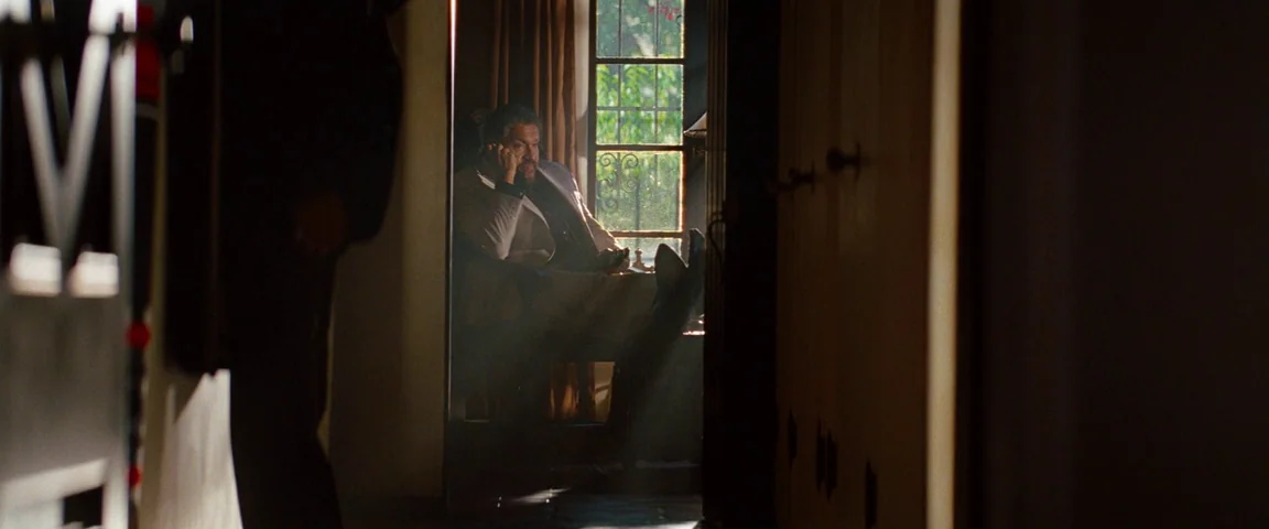 Savages (2012) movie still: medium — A man sits in a dark, shadowed room with light pouring from a window. He rests his legs on a surface and…