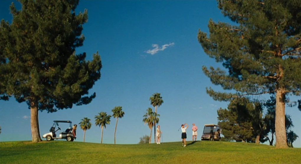The Savages (2007) movie still: wide — A manicured golf course fairway features rolling hills framed by tall pine trees. Three people stand near two…