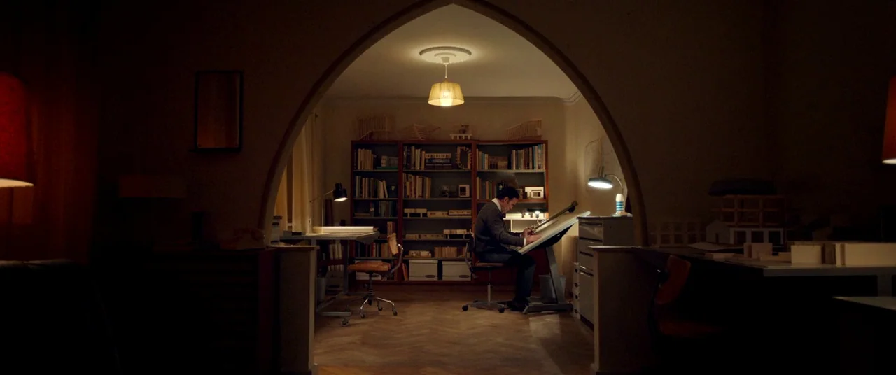 The House That Jack Built (2018) movie still: wide — A dimly lit room features a central arched doorway leading to a workspace area. A man sits at a…