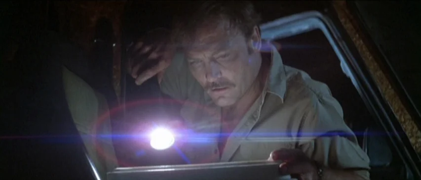 Roadgames (1981) movie still: medium — A man sits inside a dark vehicle illuminated by a single flashlight source. He holds the light over an object…