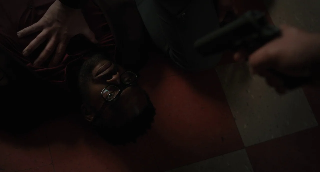 Paterson (2016) movie still: medium — A dark room features a checkered red and white tile floor. A man in eyeglasses lies on the floor while a gun…