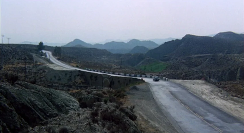 The Passenger (1975) movie still: wide — An asphalt highway winds through a desolate mountainous desert region under a hazy sky. A lone car travels…