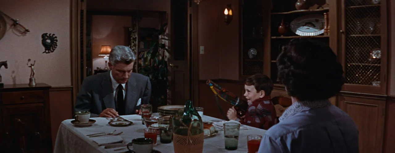 Rebel Without A Cause (1955) movie still: medium — A 1950s dining room features a set table with glass dishware and wood cabinets. A man sits at the…