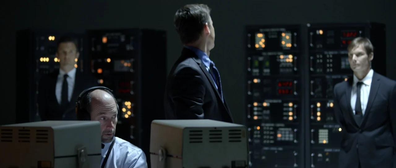 La presencia (2012) movie still: medium — A dark tech room features tall black server racks with illuminated display panels. Four men in suits…