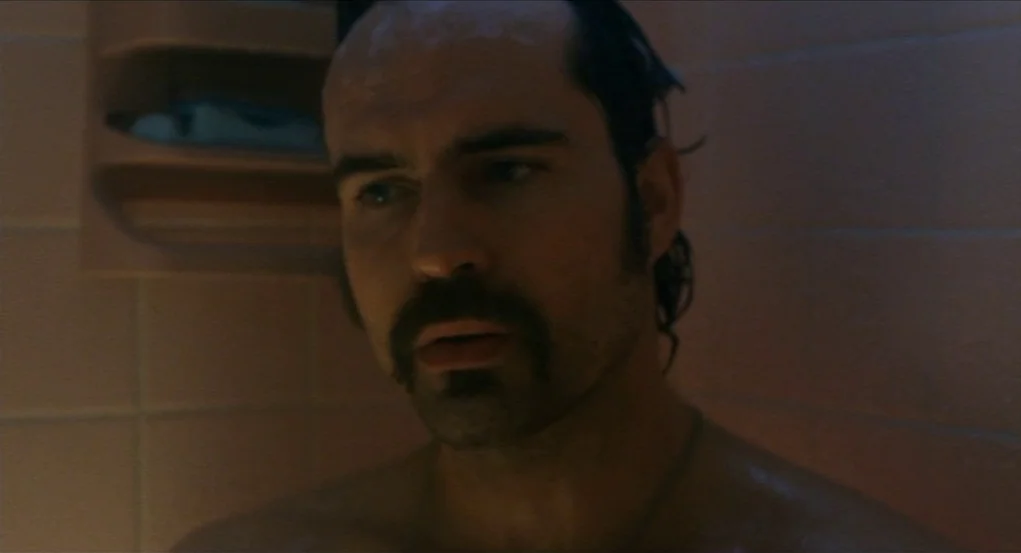 Narc (2002) movie still: closeup — A tiled stall with a mounted soap dish in a dimly lit space. A man with wet hair and a mustache looks toward the…