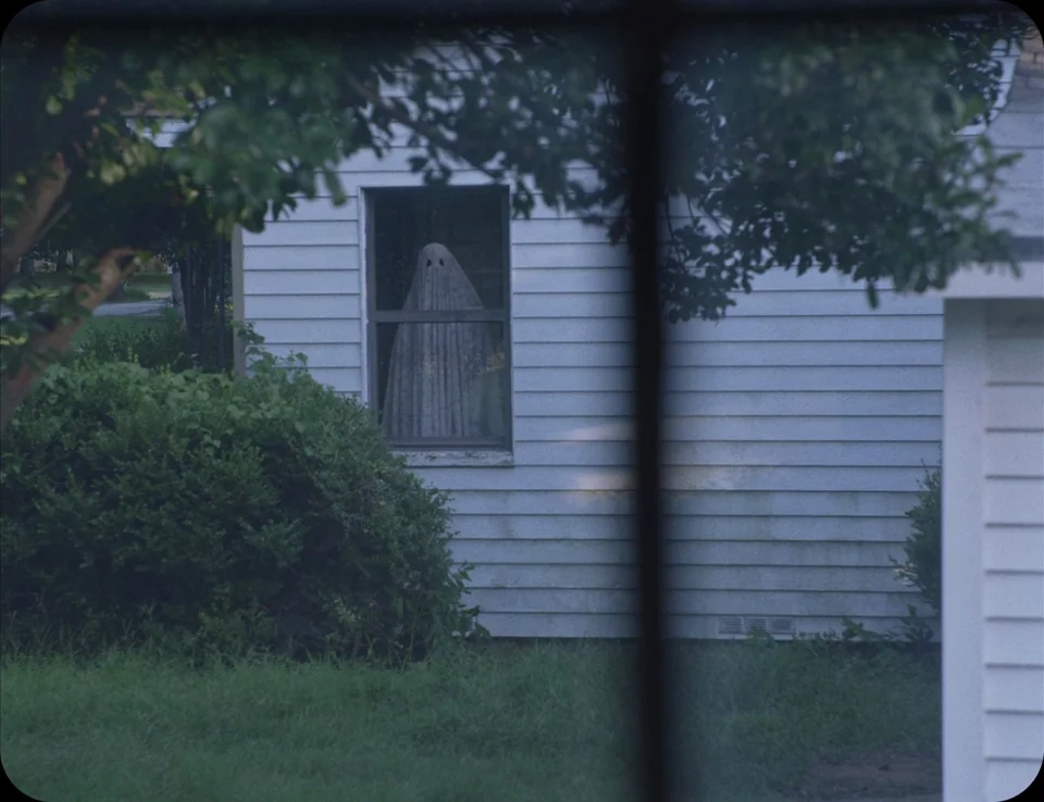 A Ghost Story (2017) movie still: wide — A suburban house exterior features light-colored horizontal siding and a single visible window. A figure…
