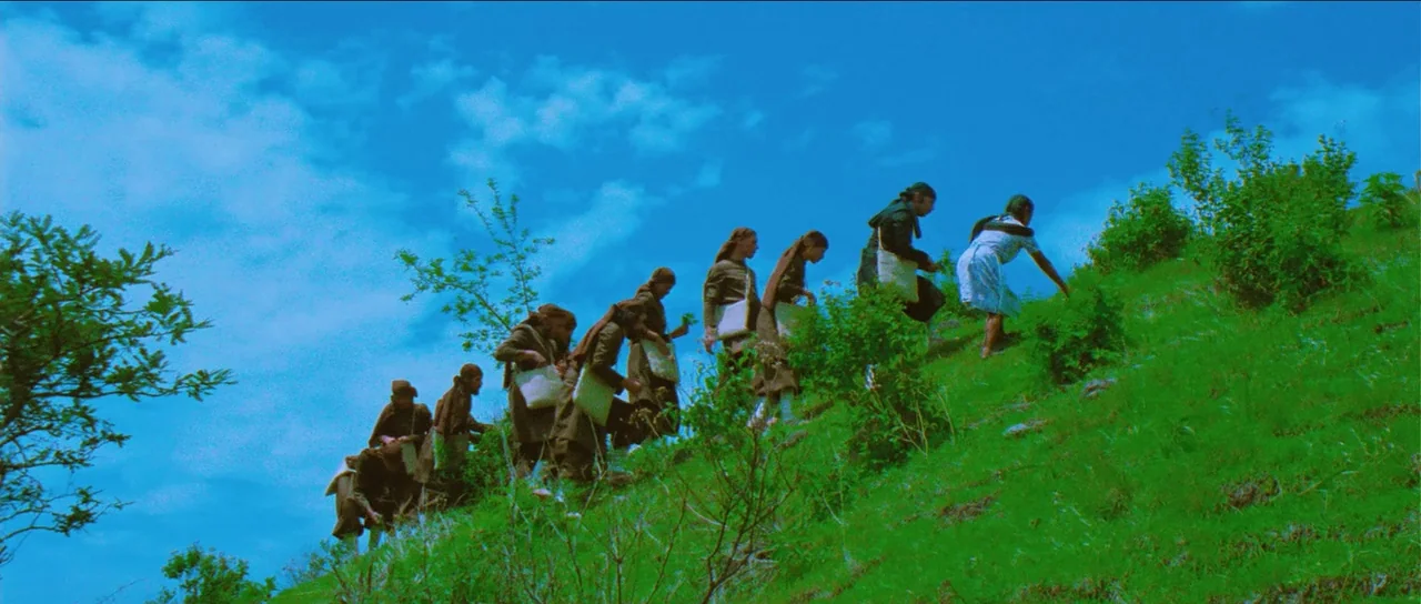 The Holy Mountain (1973) movie still: wide — A group of people climbs a steep green hillside under a clear blue sky. The individuals walk in a line…